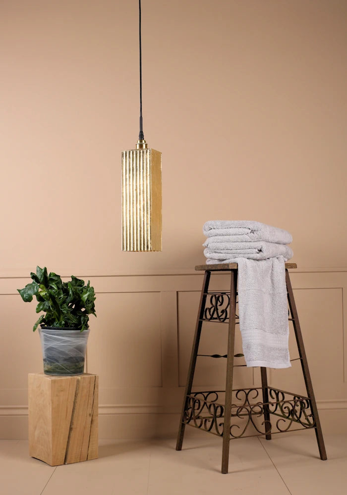 Long rectangular gold leaf bathroom pendant light switched off with bath towels and plant.