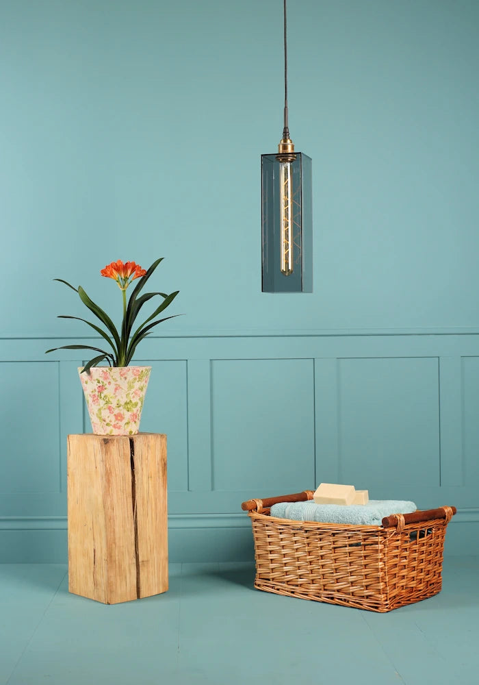 Elongated rectangular smoked glass bathroom pendant light switched off, plant, wooden basket and bath towels.