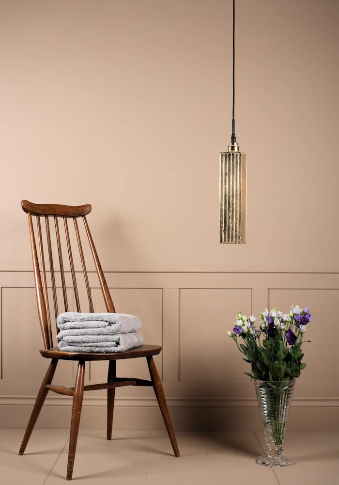 Long rectangular gold leaf bathroom pendant light switched off with bath towels on wooden chair and vase of flowers.