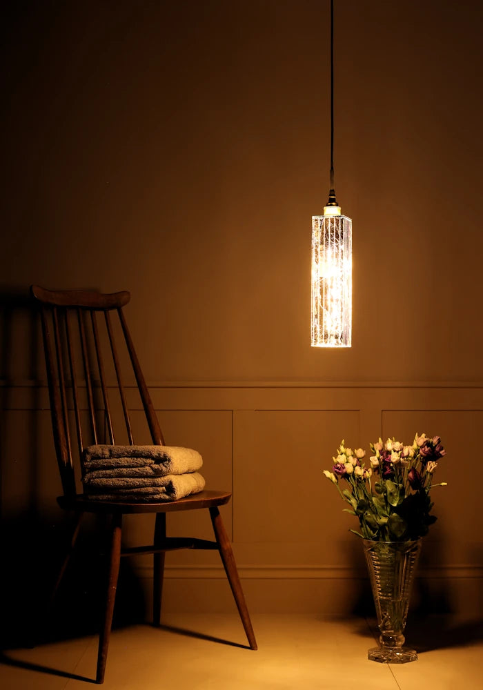 Long rectangular gold leaf bathroom pendant light casting shadows onto bath towels on wooden chair and vase of flowers.