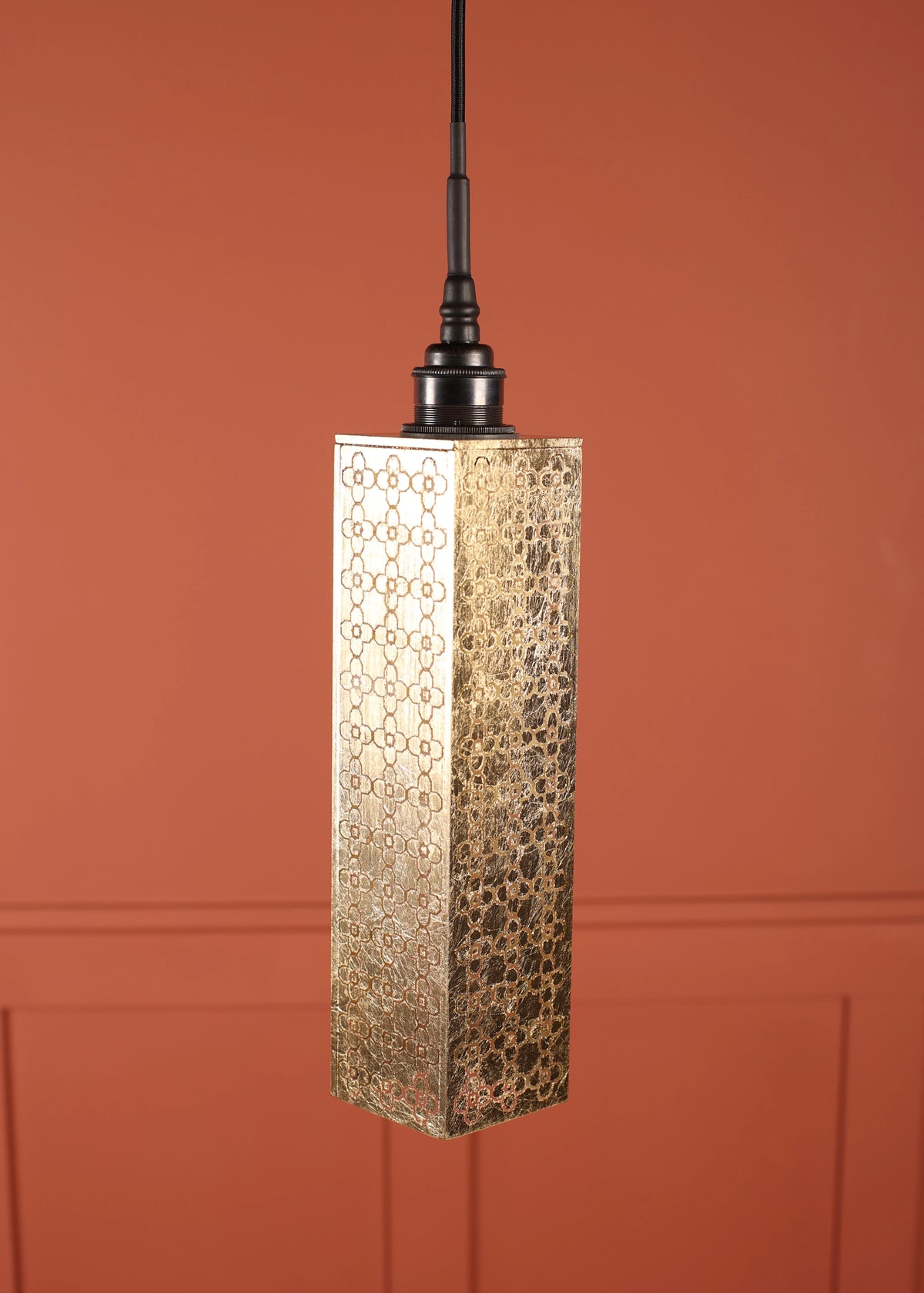 A rectangular pendant light with a hammered gold metal shade featuring a geometric cutout pattern, suspended by a black cord against a terracotta-colored paneled wall.