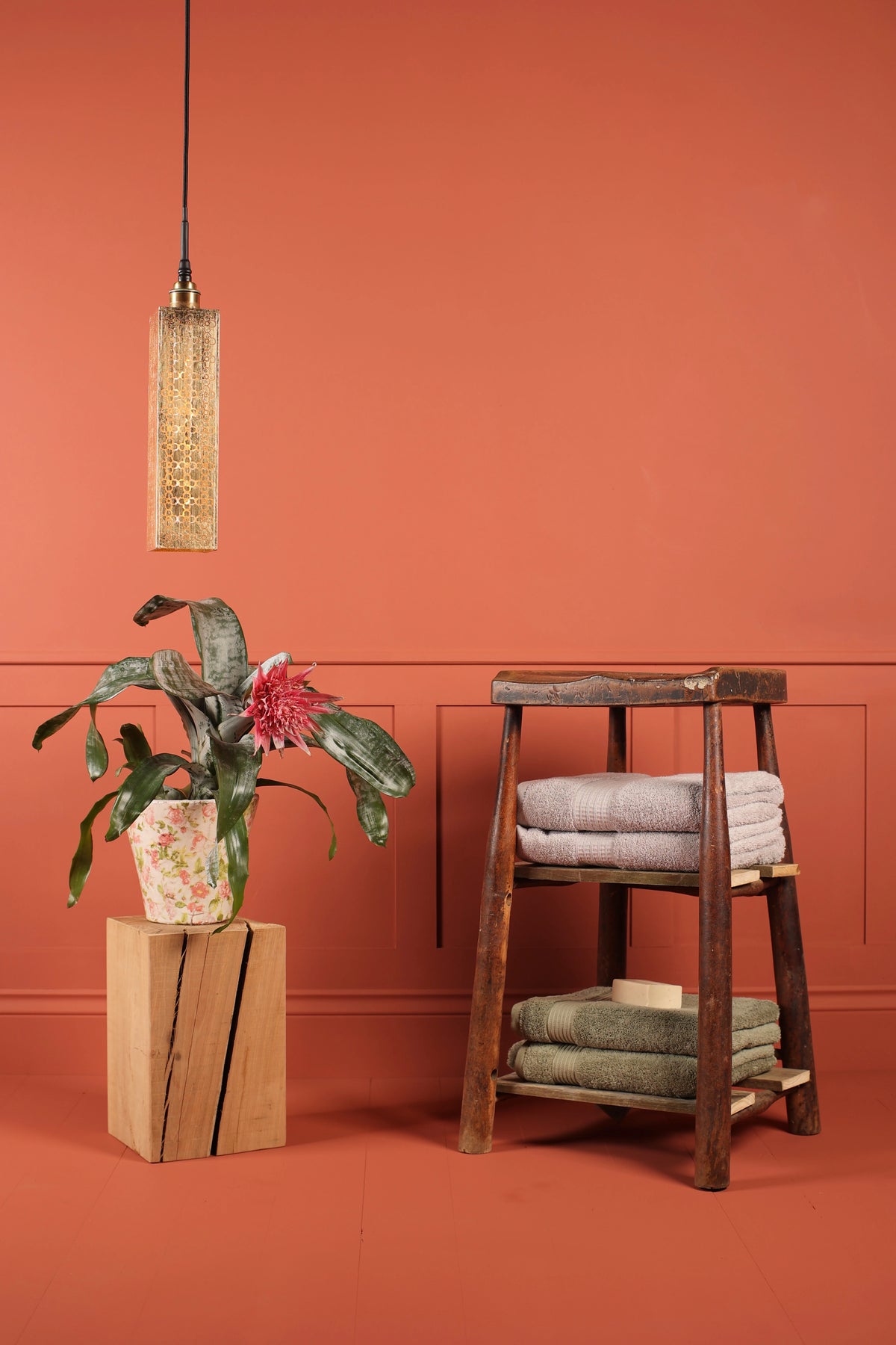 A rustic three-tier wooden stool holding folded towels, styled in a bathroom or spa setting against a terracotta orange wall with a hanging gold leaf bathroom pendant light and a potted plant on a wooden block.
