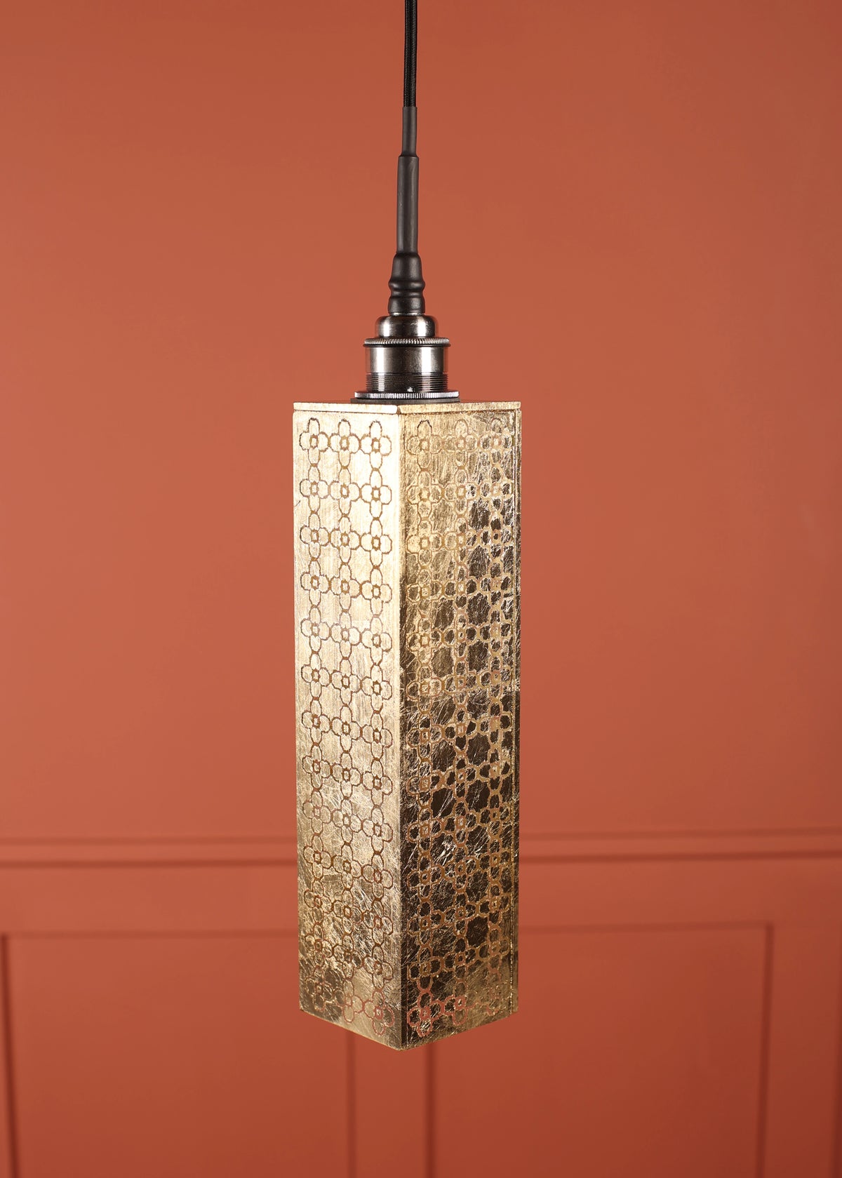 A rectangular pendant light with a hammered gold metal shade featuring a geometric cutout pattern, suspended against a terracotta-colored paneled wall.